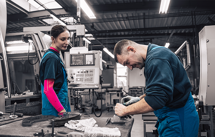 Two employees in blue work clothes are working at a table next to a machine in a production environment.