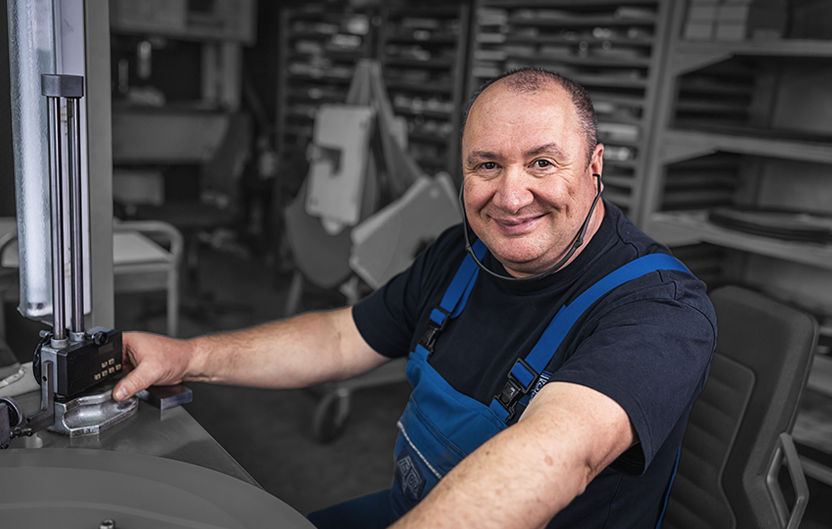 Employee in blue work clothes sits at a machine and looks into the camera.
