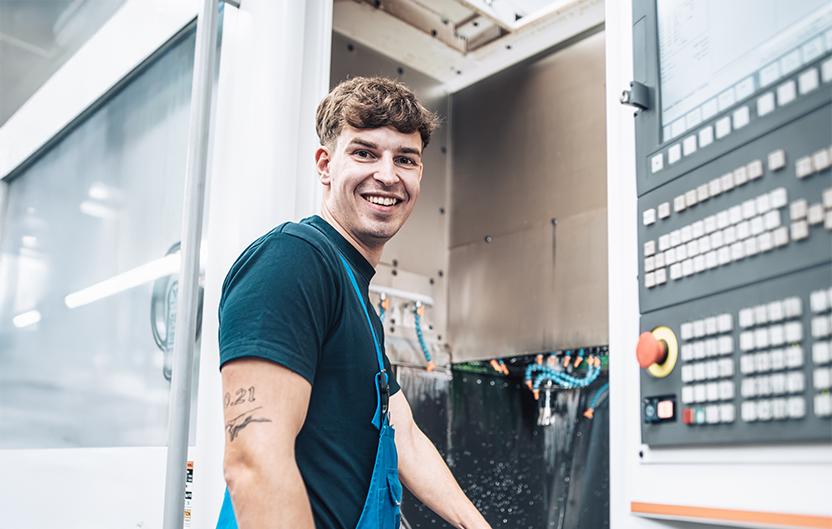 Employee in blue work clothes smiles at the camera while standing at a machine.